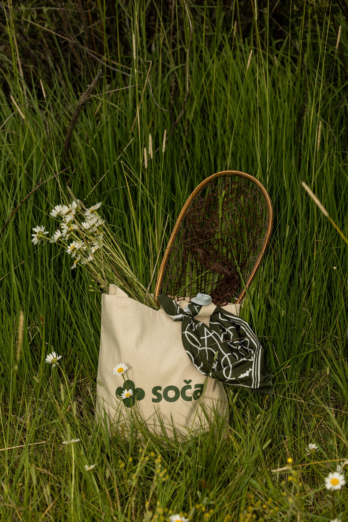 Tote bag with 'Soča' branding in a grassy field with flowers and fishing net.