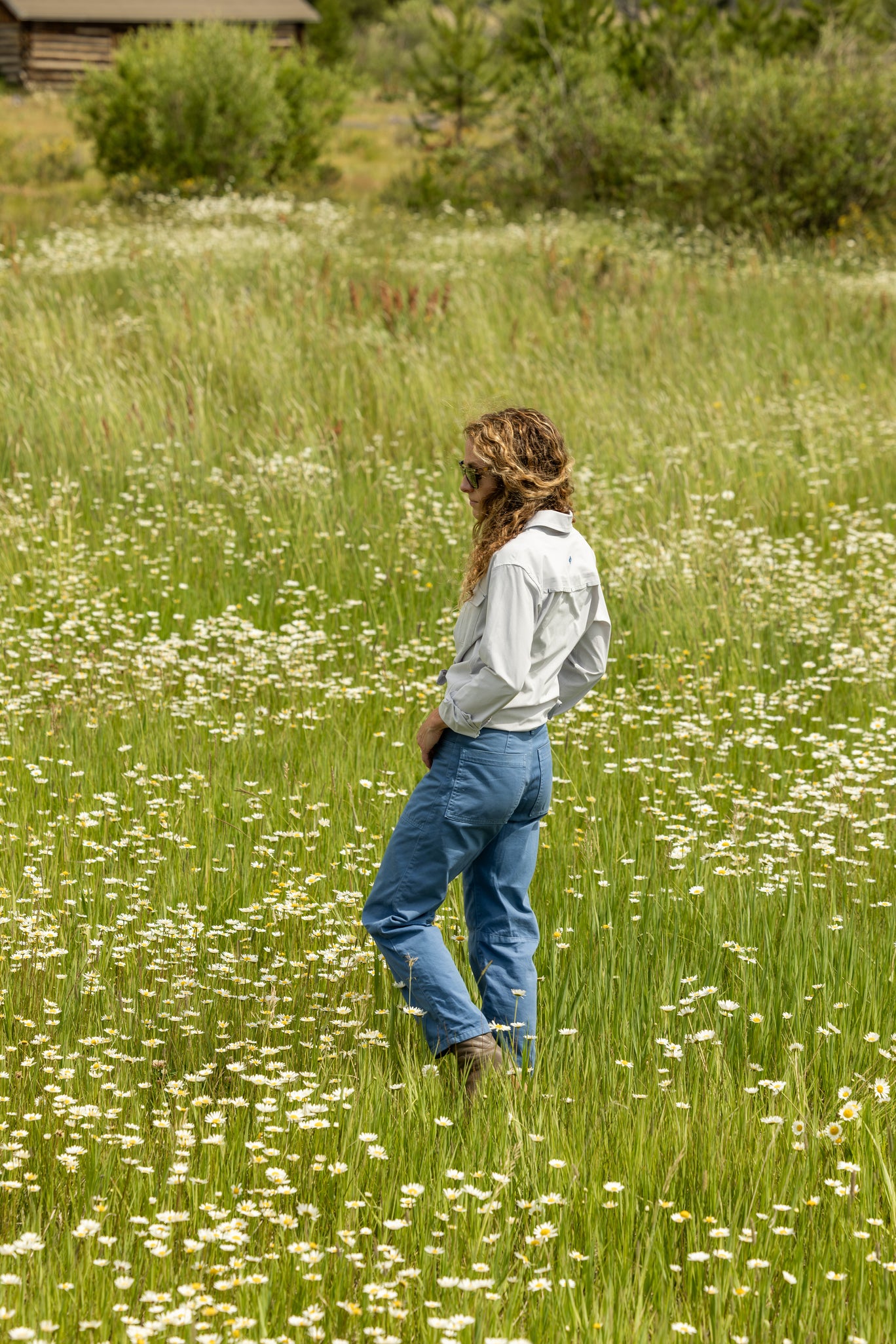 Woman walking through a field of wildflowers wearing a fly fishing button down and blue pants.
