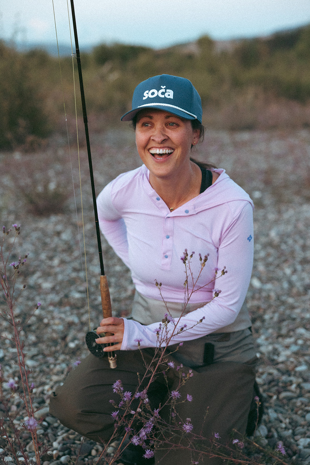 Person wearing a pink shirt and blue cap with 'soča' logo, holding a fishing rod outdoors.