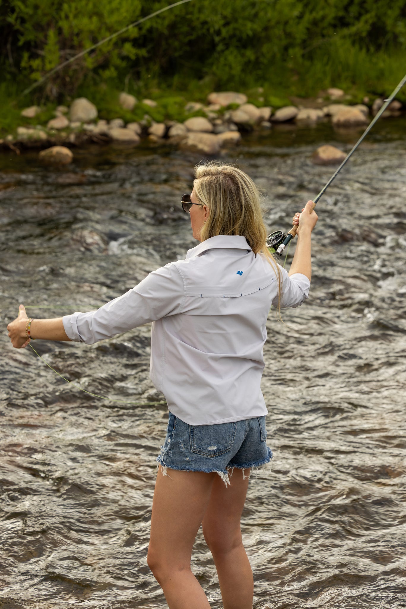 Woman fly fishing by a river with rocks and greenery in the background wearing soča fly fishing shirt