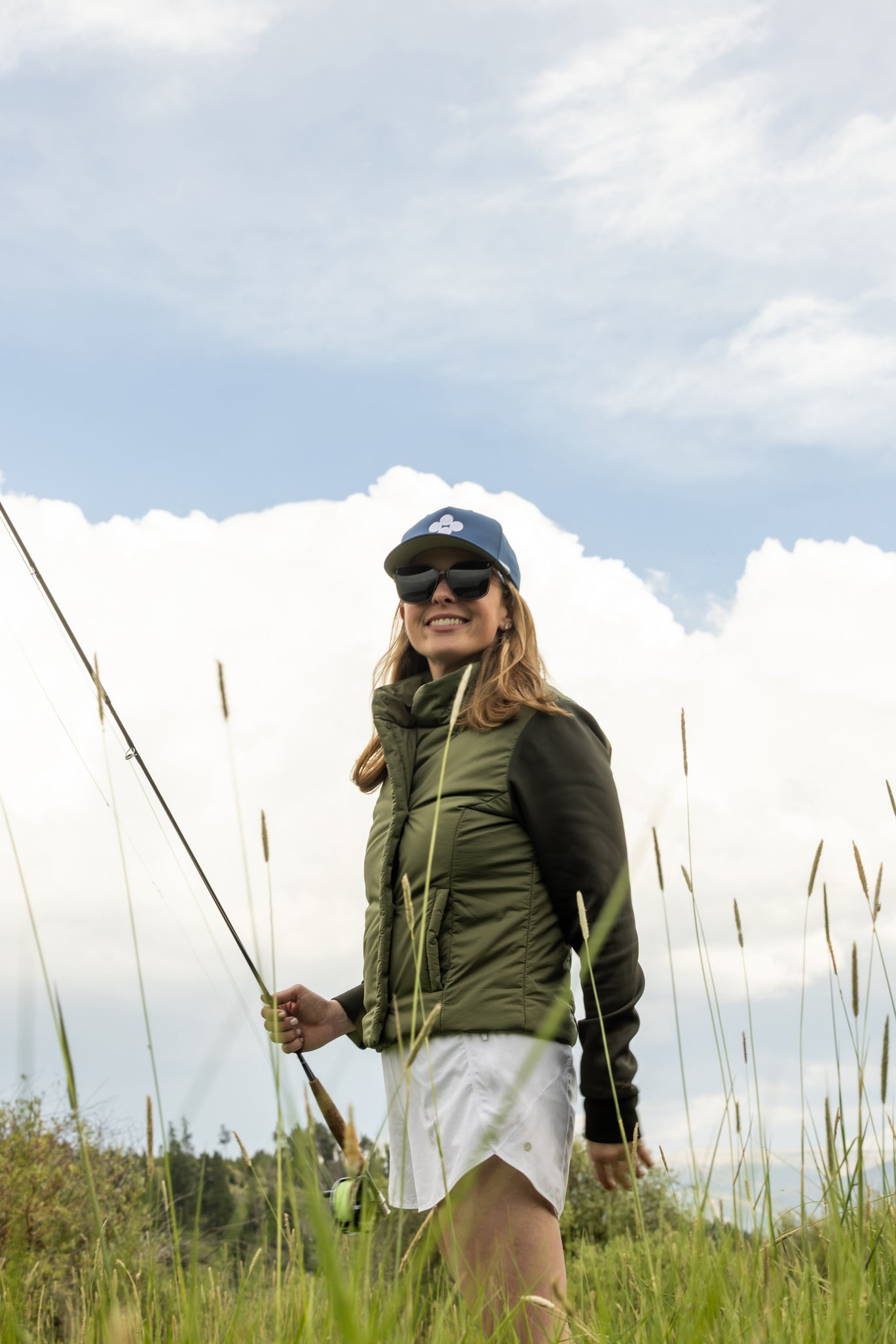 Woman in outdoor setting with fishing rod and cap, standing in grass with blue sky and clouds wearing blue hat and olive green jacket.