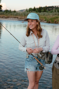 Woman fishing by in a river with a scenic background wearing blue hat and button down