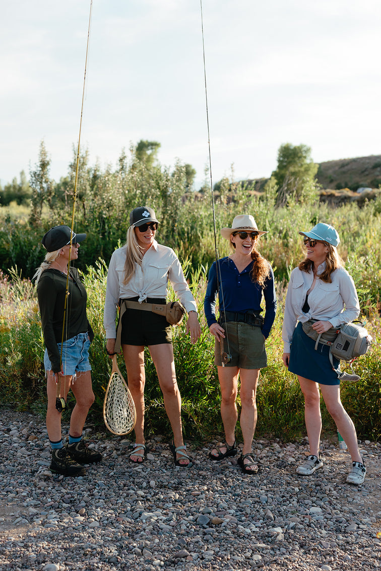 Four women standing outdoors with fishing gear, wearing hats and sunglasses.
