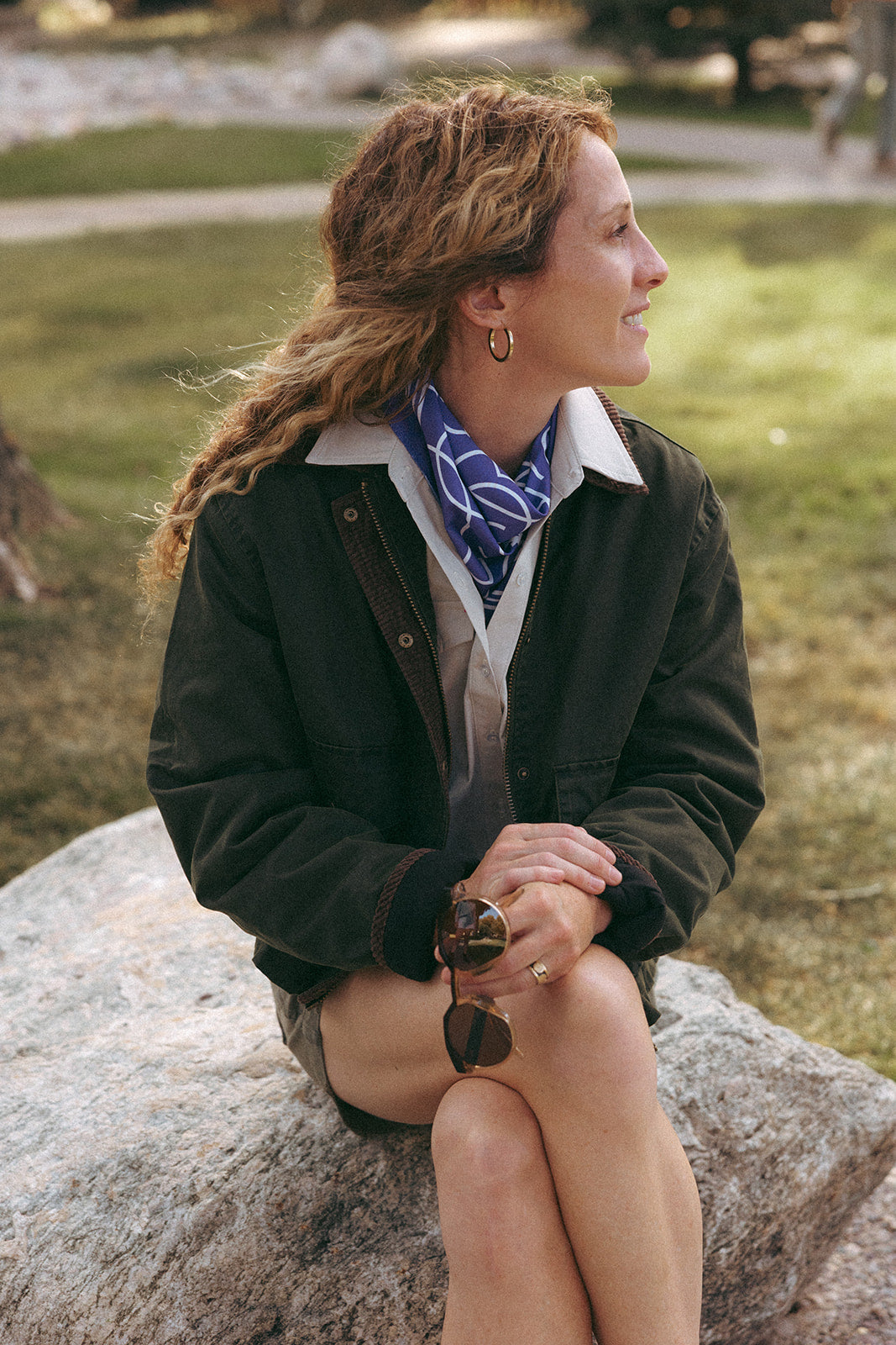 Woman sitting on a rock outdoors wearing a black jacket and purple bandana.
