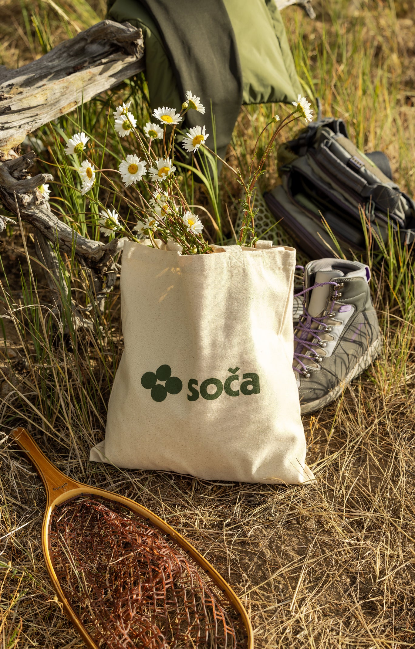 Tote bag with 'soča' logo in a natural setting with olive green jacket fishing rod and wading boots.