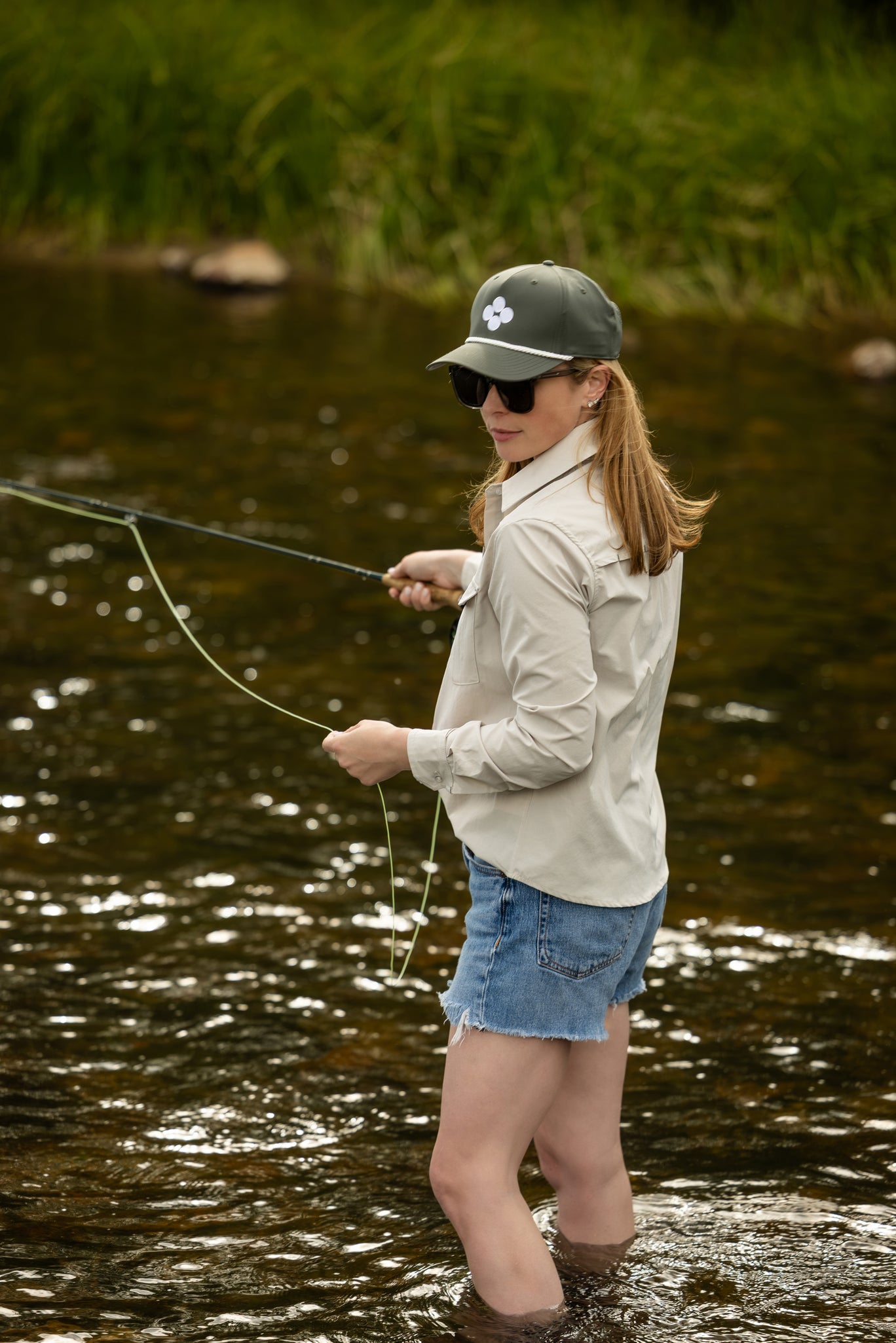 Woman fly fishing in a natural setting with water and greenery wearing olive green soča four stones logo hat and tan button down
