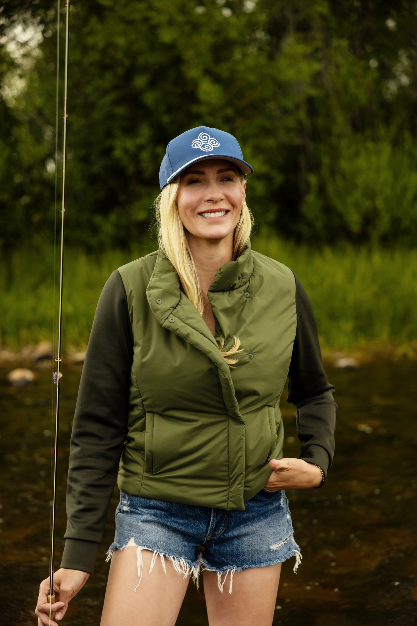 Woman wearing an olive green jacket and soča swirl logo blue cap standing outdoors by a body of water with a fishing rod.