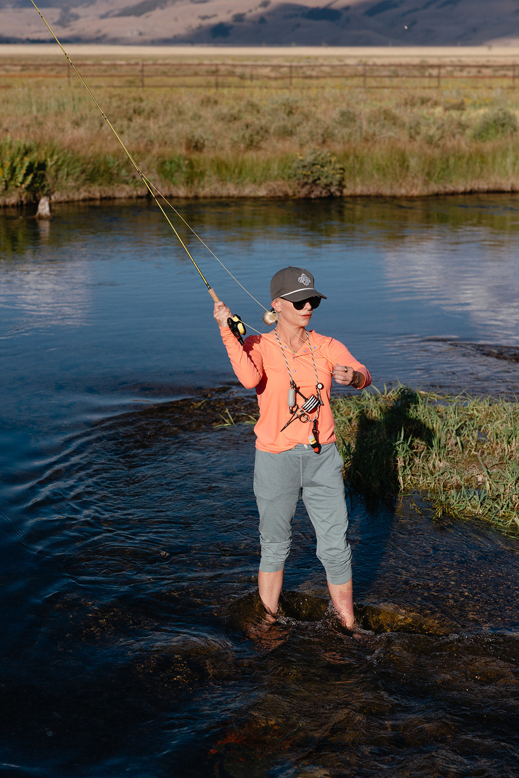 woman in fly fishing shirt in a river with mountains in the background